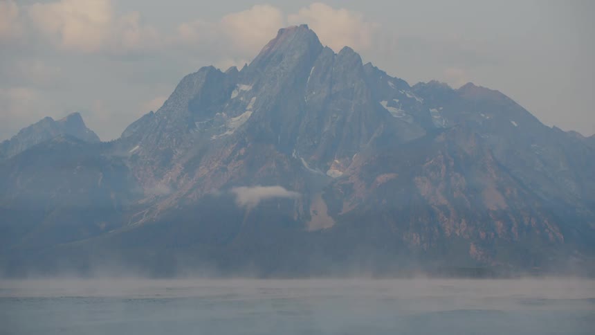 A mountain peak rises above a misty landscape its snow capped summit shrouded in clouds with a body of water stretching out in the foreground