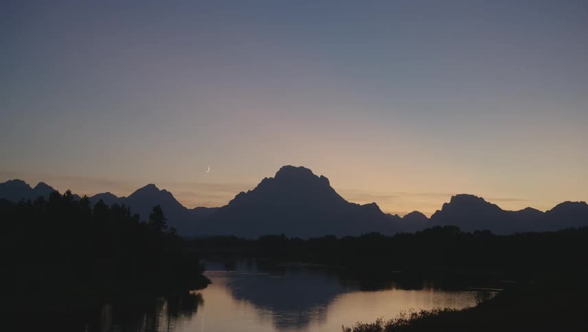 The setting sun casts a warm glow on the horizon illuminating the silhouette of a mountain range reflected in the still waters of a lake