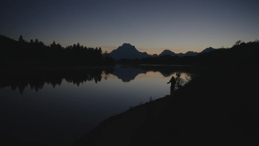 A lone fisherman casts a line into the still waters of a lake at dusk silhouetted against a backdrop of mountains and a darkening sky