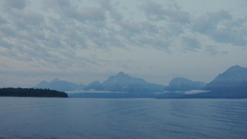 A serene scene of a lake stretching towards a distant mountain range shrouded in a veil of clouds