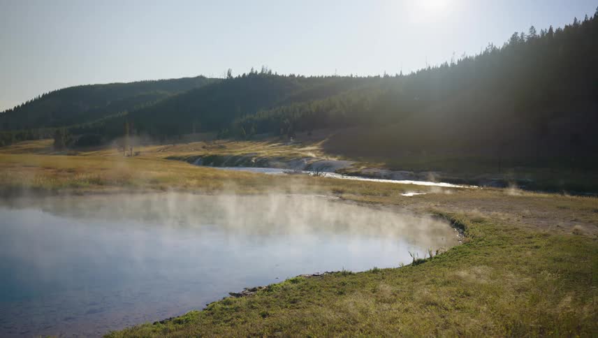 A steamy geothermal pool nestled in a valley beneath forested hills on a sunny day