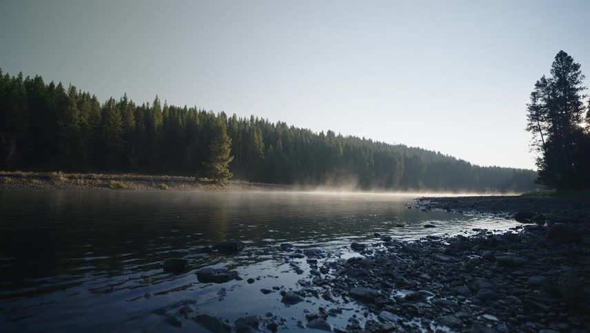 A wide shot shows a calm river with mist rising above the water framed by a rocky shore in the foreground and a dense forest in the background