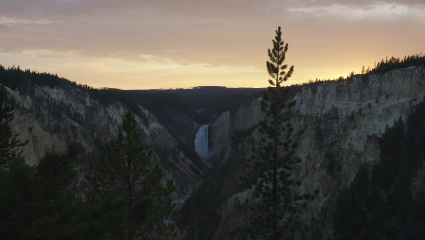The sun sets over a dramatic canyon casting a golden glow on the distant cliffs and silhouetting the trees in the foreground while a waterfall cascades down the canyon wall in the distance