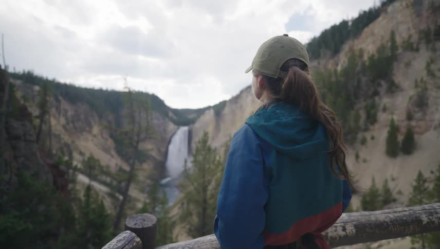 A woman walks up to a railing overlooking a waterfall in a canyon and takes a photograph