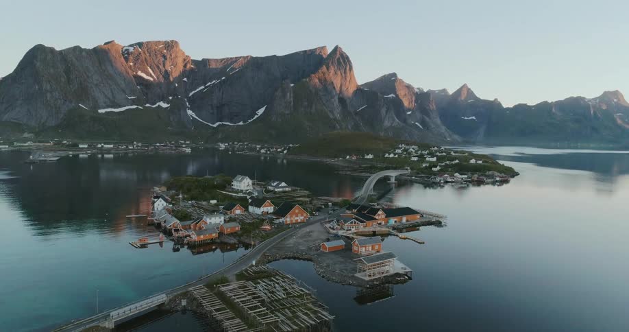 An aerial view of a coastal village with colorful houses and a bridge connecting it to the mainland nestled between dramatic mountains at sunset in lofoten norway