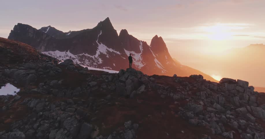 A person stands on a rocky outcrop overlooking a dramatic mountain range with snow patches bathed in the warm light of a setting or rising sun