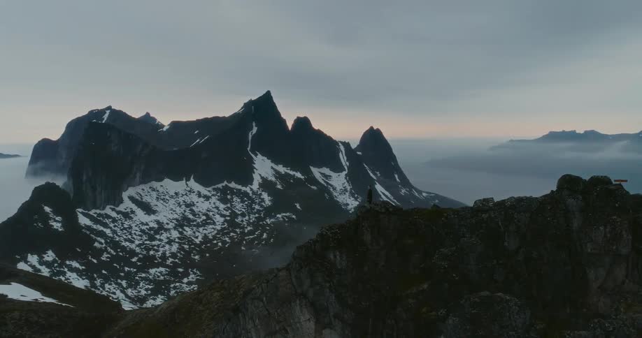 Silhouette of a person standing on a mountain range with jagged peaks partially covered in snow rises above a misty landscape
