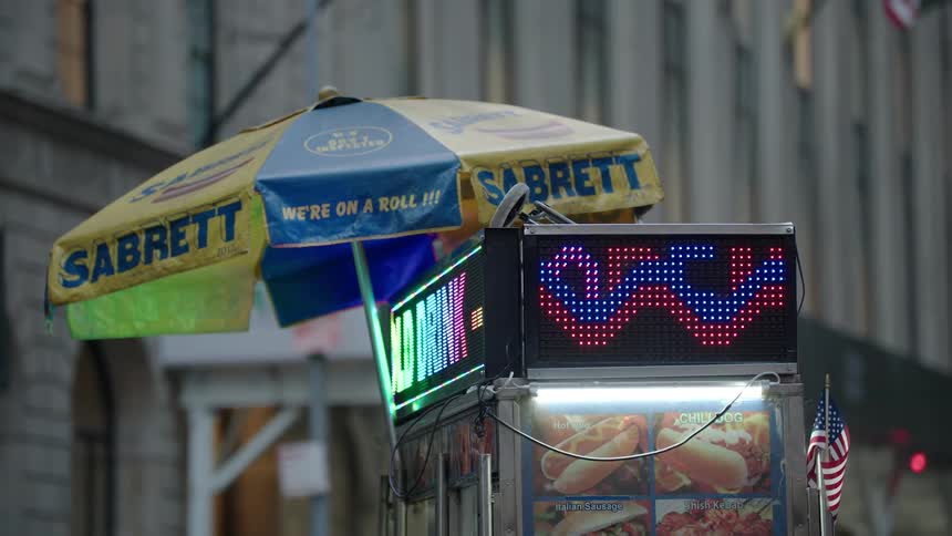A hot dog cart with a yellow umbrella and a digital sign stands on a city street