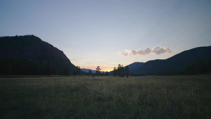A person walking on a grassy field flanked by silhouetted mountains at sunset