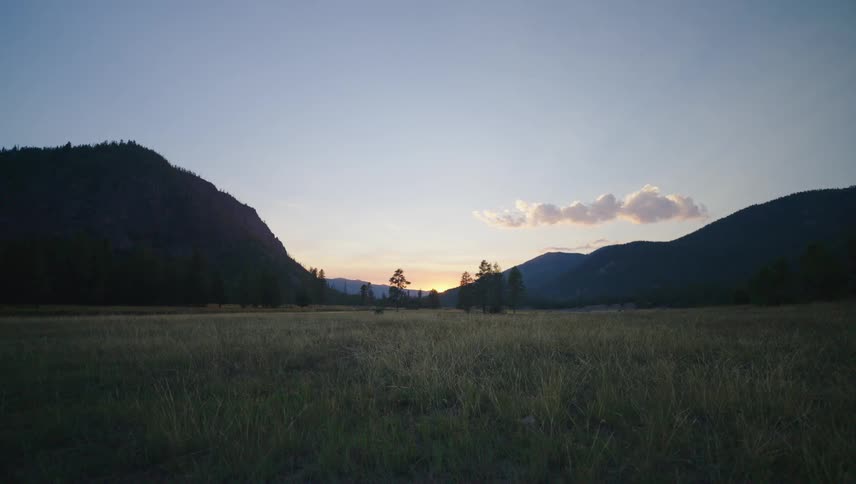 A vast grassy plain stretches towards a distant mountain range as the sun sets casting long shadows across the landscape and illuminating the clouds with a warm glow