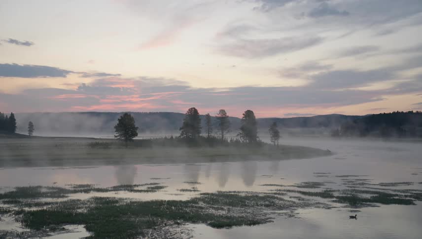 Landscape of a lake surrounded by mountains and vegetation on a foggy dawn