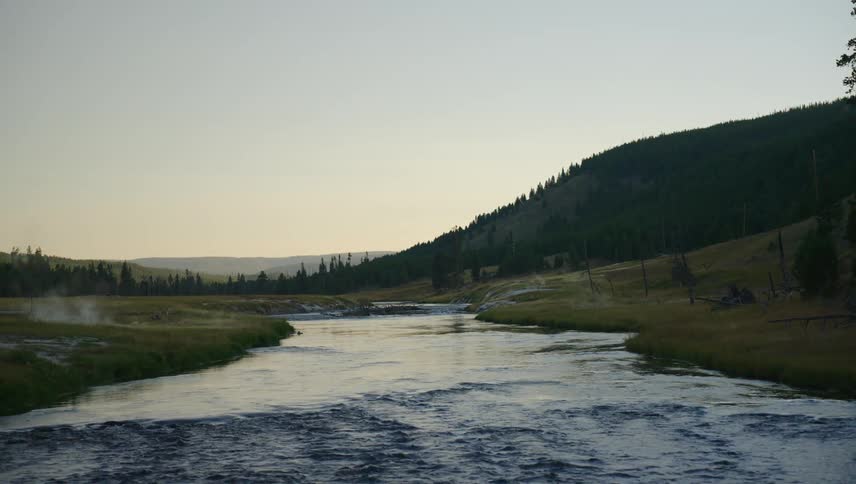 A serene river flows through a valley flanked by grassy banks and forested hills under a clear sky