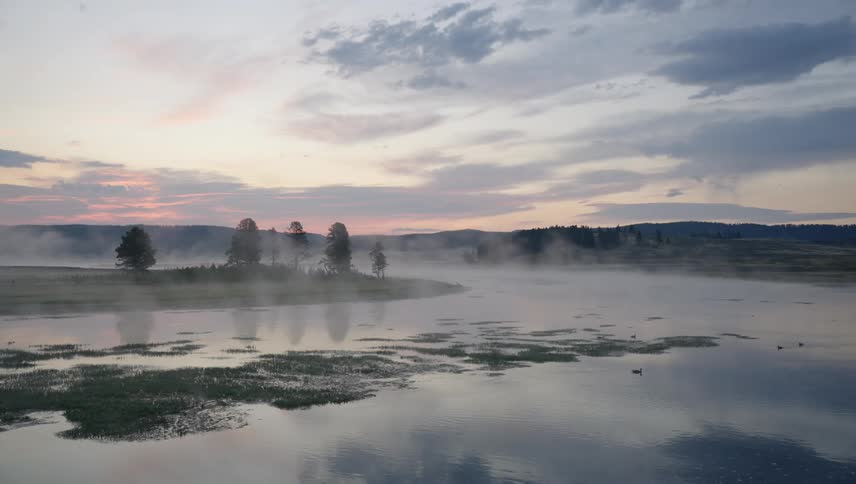 A serene landscape features a mistcovered lake reflecting the pastel hues of a sunrise or sunset under a partly cloudy sky
