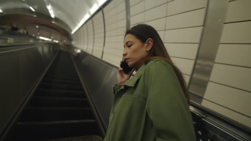 A woman rides an escalator up at a subway station while talking on a phone