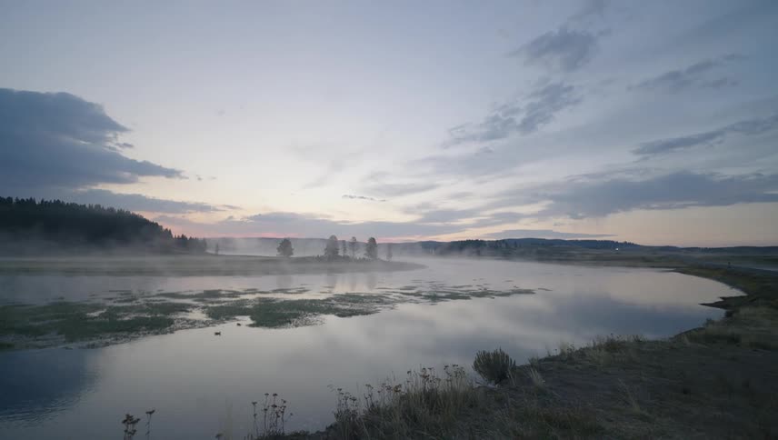 Fog flows over a river near a forest while red tints the horizon