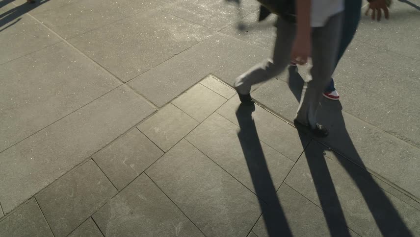 A busy sidewalk is filled with people walking their shadows stretching across the pavement