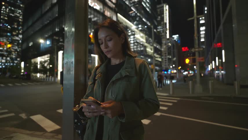 A woman stands on the corner of a city street at night texting on her phone