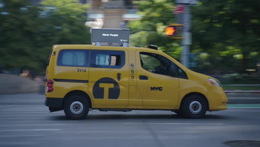 A yellow taxi van drives along a busy street in a downtown metropolis