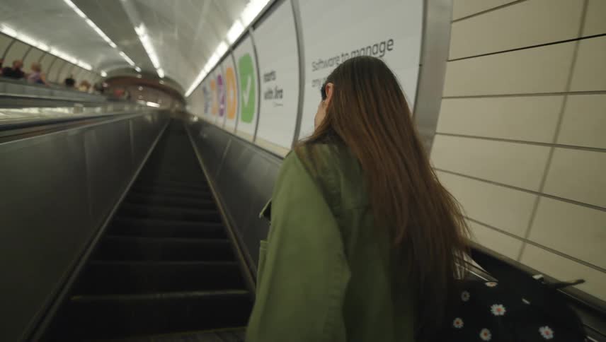 A woman in a green coat is talking on the phone while going up on an escalator and she is smiling