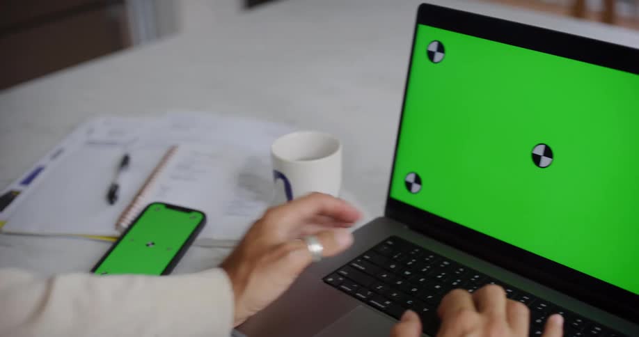 A man wearing rings and a white sweatshirt types on a laptop and then uses a smartphone lying nearby