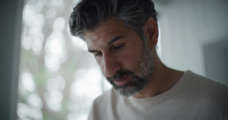 A bearded man with a sweatshirt looks down on his device in the kitchen