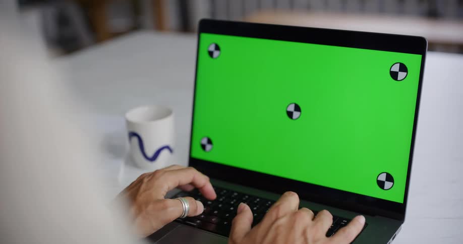 A persons hands are shown typing on a laptop with a green screen while a mug sits on the table beside it