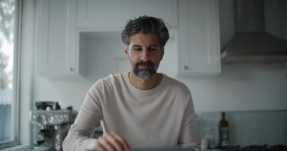 A bearded man in a cream sweatshirt ends a video call on his laptop and then picks up his smartphone to use in the kitchen
