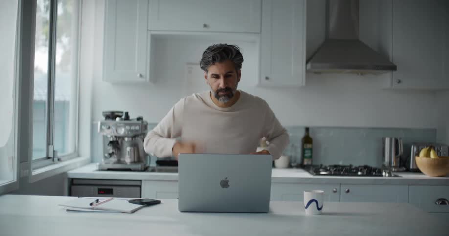 A bearded man wearing a cream sweatshirt spruces himself up to look more professional before his video call on the laptop at the kitchen island