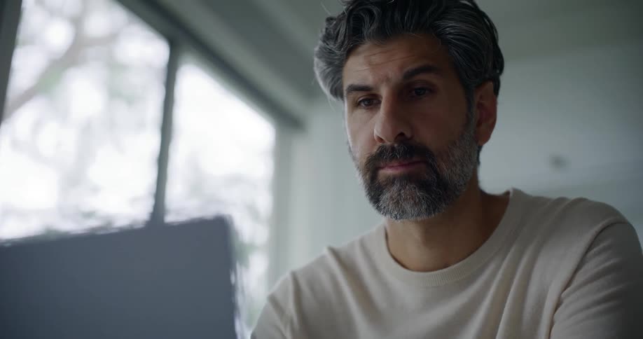 A bearded man in a cream sweatshirt uses a laptop in the kitchen