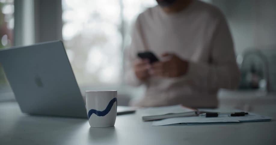 Bearded man in a cream shirt uses smartphone and reaches out to the coffee cup near the laptop at the kitchen island