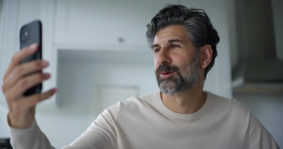 Bearded man wearing a cream sweatshirt has a video call on the smartphone in the kitchen