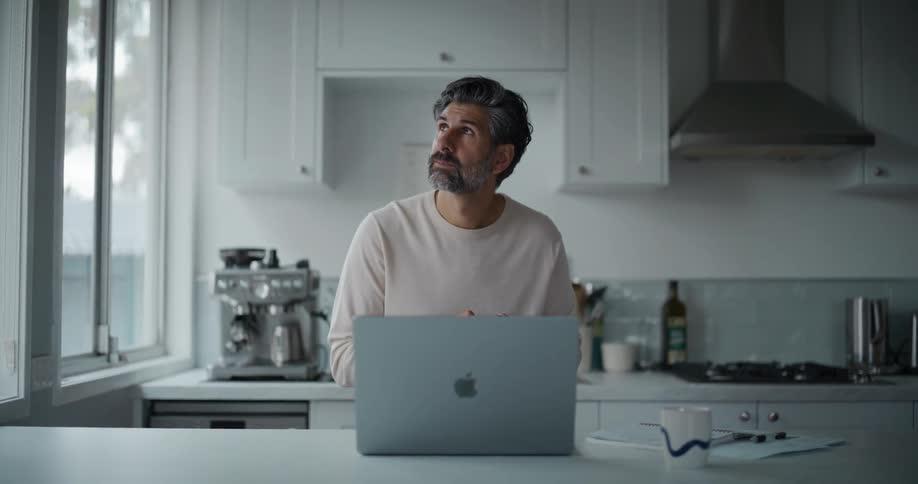A man stands in his kitchen while looking out the window and scrolling on his smartphone