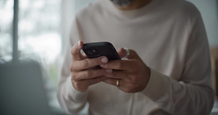 Older man uses a smartphone while sitting near a computer during the day