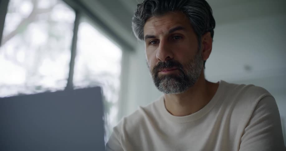 Bearded man in a cream sweatshirt uses laptop in the kitchen