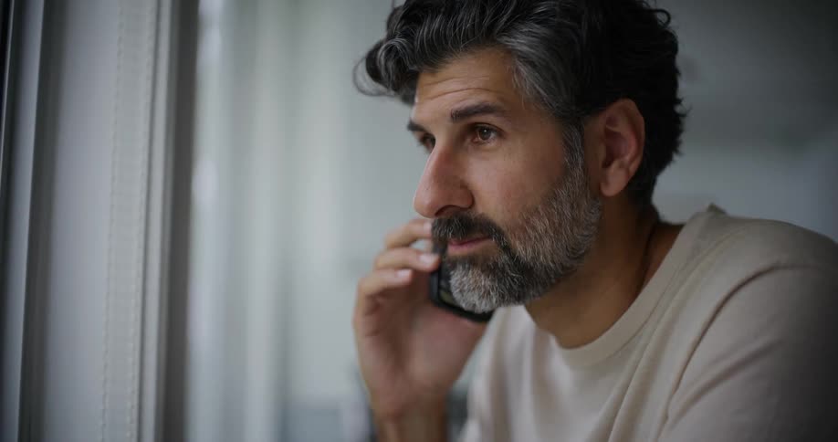 A bearded man wearing a cream shirt talks on the smartphone while looking out the window