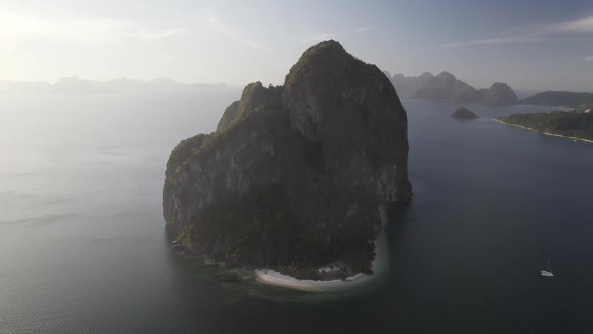 A solitary rocky island with a small sandy beach rises from the calm ocean surrounded by hazy mountains in the distance