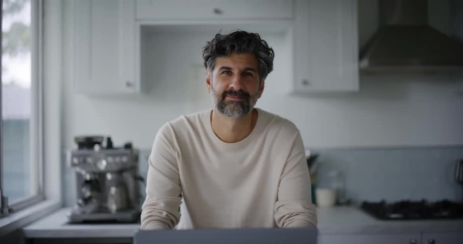 A bearded man in a white sweatshirt uses a laptop and looks up at the camera smiling in the kitchen