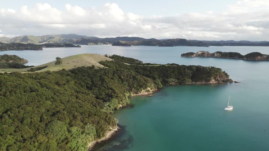 A sailboat glides through a calm bay surrounded by lush green islands and hills under a clear blue sky dotted with fluffy white clouds