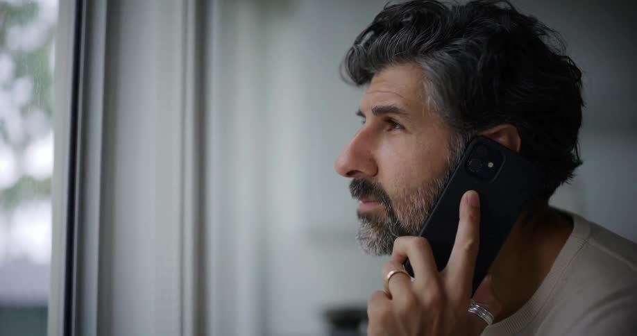 A bearded man with a cream sweatshirt talks on the smartphone