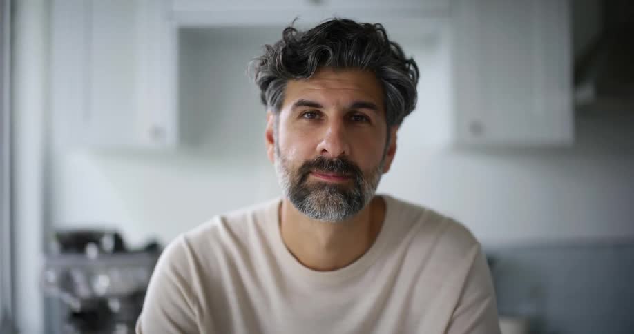 A bearded man in a white sweatshirt uses a device and looks up at the camera smiling in the kitchen