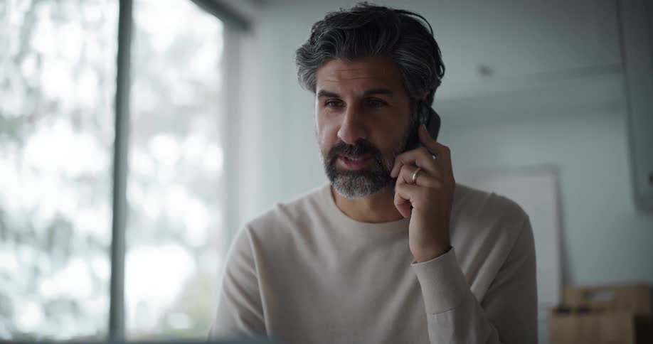 A bearded man wearing a cream sweatshirt uses a laptop and answers the phone in the kitchen