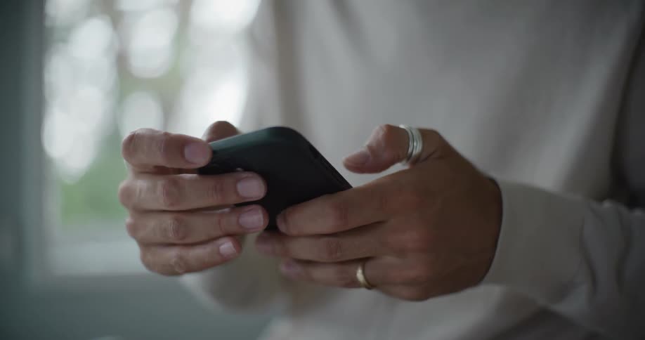 A mans hands holding a black smartphone with fingers delicately tapping the screen adorned with several rings
