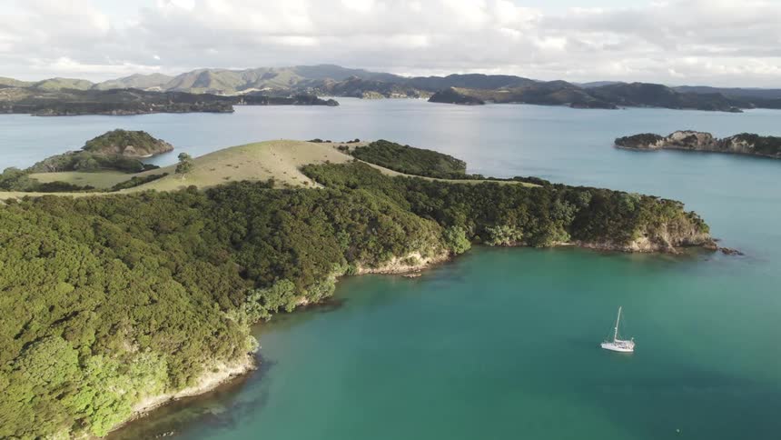 A sailboat glides through a calm bay surrounded by lush green islands and hills under a clear sky