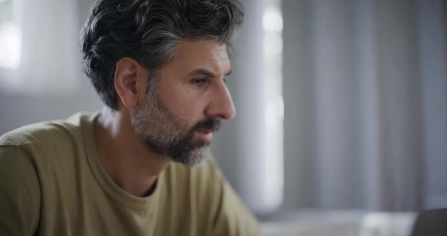 A bearded man with black and white hair is talking while there is a notebook in front of him