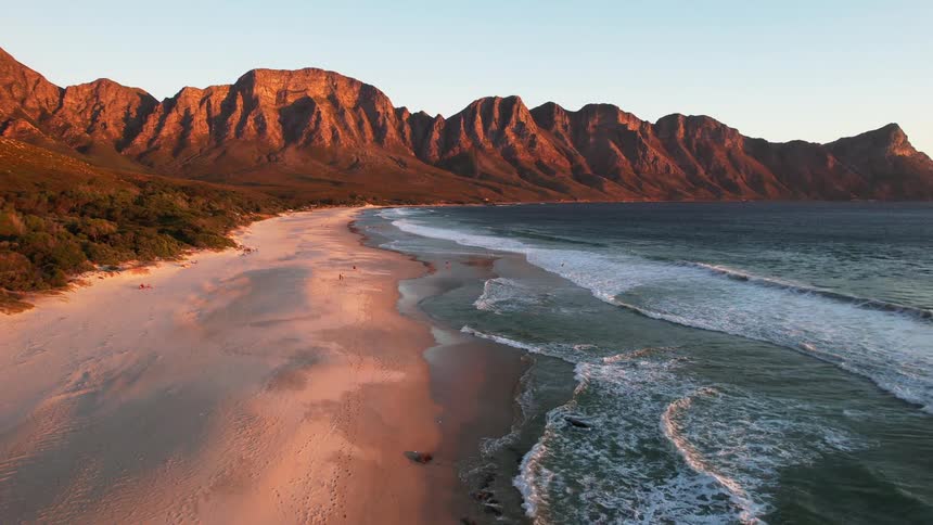 A pristine sandy beach stretches along a coastline with dramatic mountains bathed in the warm light of sunset in the background and a group of people enjoying the scenic view