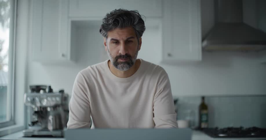 A bearded man wearing a cream sweatshirt spruces himself up before a video call on the laptop at the kitchen island