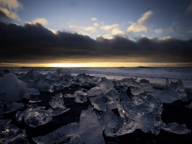 Large chunks of ice are scattered across a dark volcanic sand beach as dramatic waves crash against the shore under a stormy sky while the sun rises over the sea in the distance