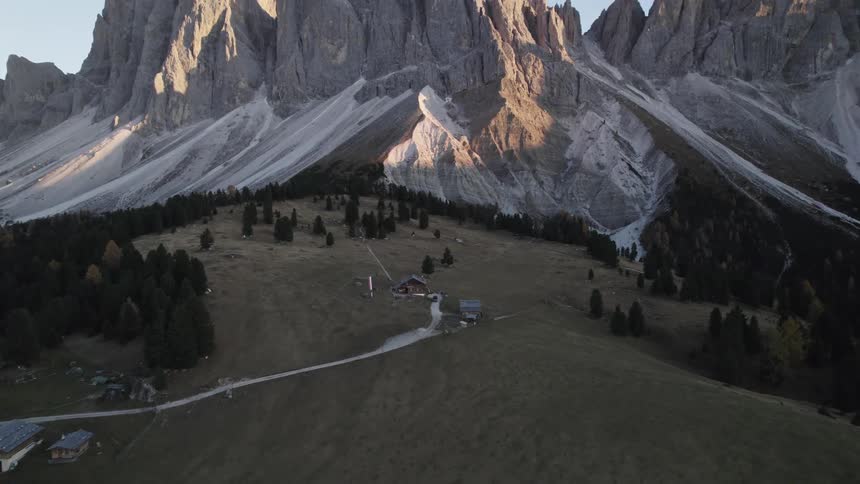 A winding dirt road leads to small buildings nestled amidst a verdant valley with dramatic jagged mountain peaks towering in the background as the sun casts long shadows across the landscape