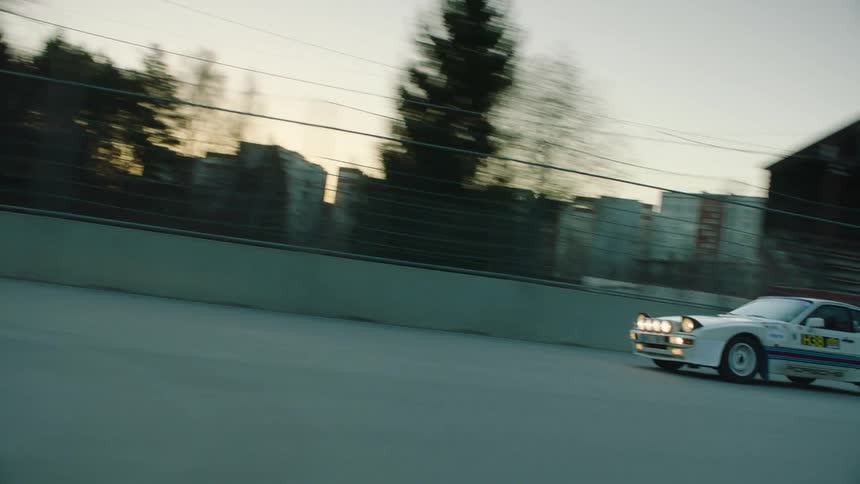 A white porsche 944 rally car with martini livery is shown racing along an outdoor track