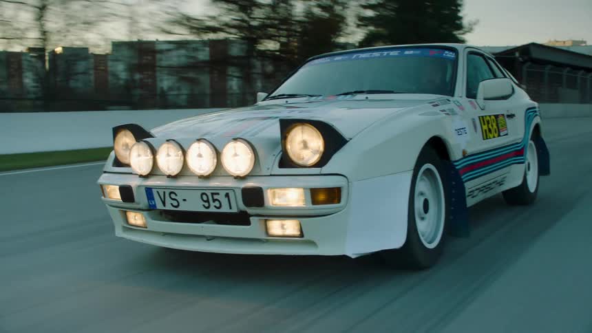 A white porsche 924 rally car with a distinctive blue red and yellow stripe and numerous headlights speeds along a track